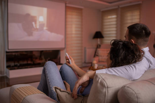 A couple relaxes on a couch, watching a movie on a home theater projector screen in a cozy living room.